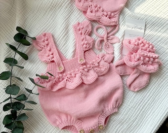 May include: A pink knitted baby outfit set with a hat, a romper, and booties. The set features a delicate crocheted design with pom-poms.
