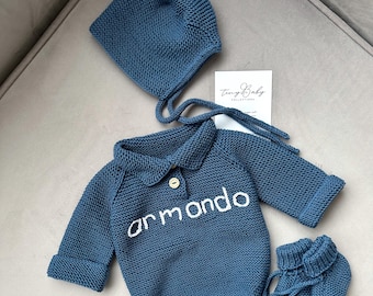 May include: A blue knitted baby outfit with a matching hat and booties. The outfit has a collared neckline and the word "armando" embroidered on the front.