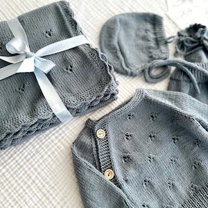 K&ouml;nnte beinhalten: Ein graues gestricktes Baby-Outfit-Set mit einem Cardigan, einer M&uuml;tze und einer Decke. Der Cardigan hat einen Knopfverschluss und die Decke ist mit einem blauen Band gebunden.