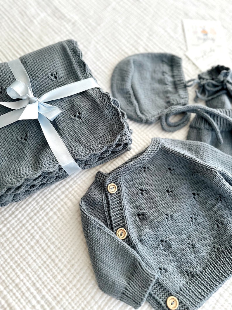 K&ouml;nnte beinhalten: Ein graues gestricktes Baby-Outfit-Set mit einem Cardigan, einer M&uuml;tze und einer Decke. Der Cardigan hat einen Knopfverschluss und die Decke ist mit einem blauen Band gebunden.