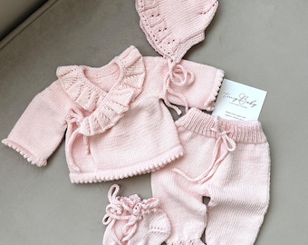 Può includere: Un completo di abbigliamento per neonati in maglia rosa chiaro. Il set comprende un cardigan avvolgente con finiture a balze, pantaloni con vita con coulisse e polsini a balze, un cappello e scarpine. È presente anche una carta con il testo "Tiny Baby Collections".