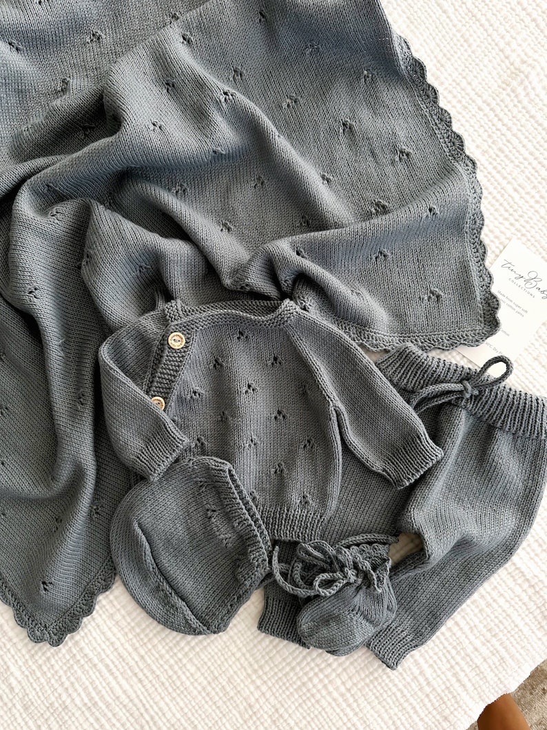 K&ouml;nnte beinhalten: Ein graues gestricktes Baby-Outfit-Set bestehend aus einem Pullover, einer Hose, einer M&uuml;tze und Stiefeln. Das Set hat ein zartes Blumenmuster und einen gewellten Rand an der Decke.