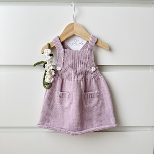 May include: A lavender knitted baby dress with two pockets and a white button detail. The dress is hanging on a wooden hanger with a white tag that says "Tiny Baby Collections".