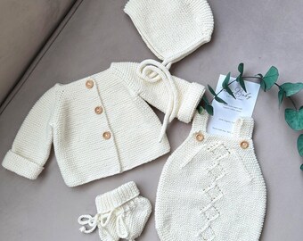 Könnte beinhalten: Ein weißes Strick-Baby-Outfit-Set bestehend aus Mütze, Cardigan, Strampler und Stiefeln. Das Set verfügt über ein zartes Zopfmuster und Knöpfe aus Holz.