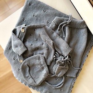 K&ouml;nnte beinhalten: Ein graues gestricktes Baby-Outfit-Set bestehend aus einem Pullover, einer Hose, einer M&uuml;tze und Stiefeln. Das Set liegt auf einer wei&szlig;en Oberfl&auml;che.