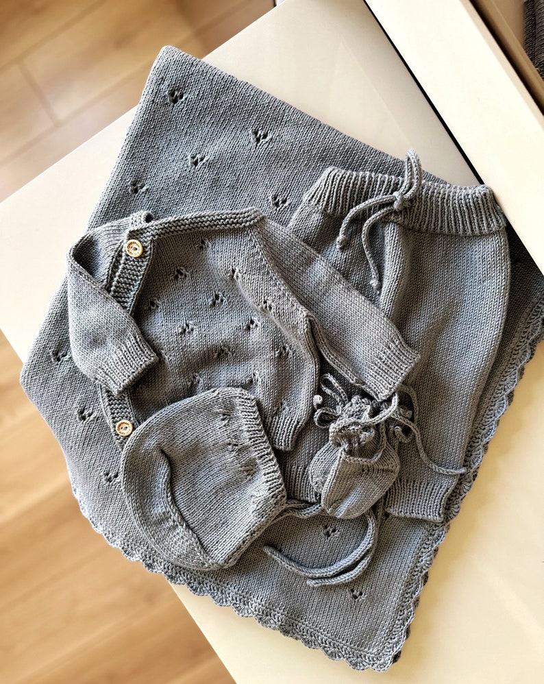 K&ouml;nnte beinhalten: Ein graues gestricktes Baby-Outfit-Set bestehend aus einem Pullover, einer Hose, einer M&uuml;tze und Stiefeln. Das Set liegt auf einer wei&szlig;en Oberfl&auml;che.