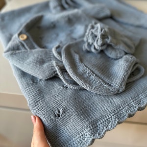 K&ouml;nnte beinhalten: Ein hellblaues gestricktes Baby-Outfit mit einem gewellten Rand. Das Outfit beinhaltet eine M&uuml;tze, einen Pullover und eine Hose.