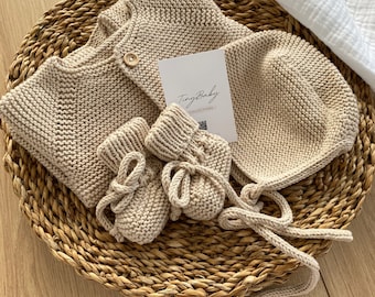 May include: A beige knitted baby cardigan, hat, and booties set. The set is laid out on a woven basket with a white muslin blanket in the background. The tag reads "Tiny Baby Collections".
