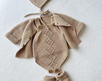 May include: A beige knitted baby outfit set, including a bonnet, cardigan, romper, and booties. The romper features a diamond pattern and wooden buttons. The set is made of soft, textured yarn, perfect for a newborn. The booties have a tie closure.