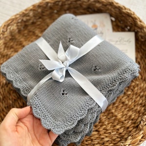 K&ouml;nnte beinhalten: Eine graue gestrickte Babydecke mit einem gewellten Rand, gebunden mit einem wei&szlig;en Satinband. Die Decke ist gefaltet und in einem geflochtenen Korb gelegt.