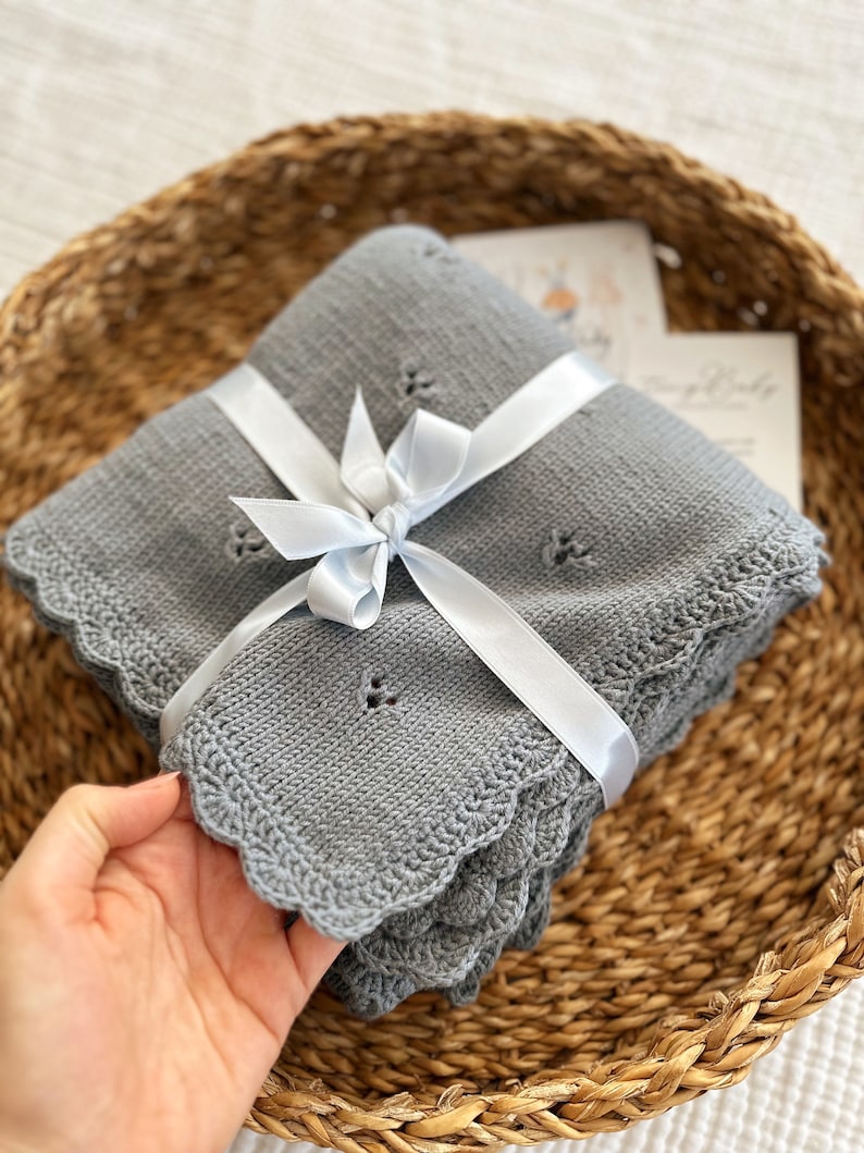 K&ouml;nnte beinhalten: Eine graue gestrickte Babydecke mit einem gewellten Rand, gebunden mit einem wei&szlig;en Satinband. Die Decke ist gefaltet und in einem geflochtenen Korb gelegt.