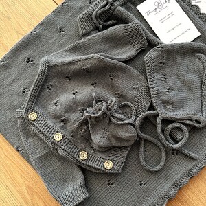 K&ouml;nnte beinhalten: Ein graues gestricktes Baby-Outfit-Set mit Cardigan, M&uuml;tze und Booties. Das Set verf&uuml;gt &uuml;ber ein zartes Lochmuster und Holzkn&ouml;pfe. Das Outfit ist aus 100% Bio-Baumwolle gefertigt und nur handwaschbar.