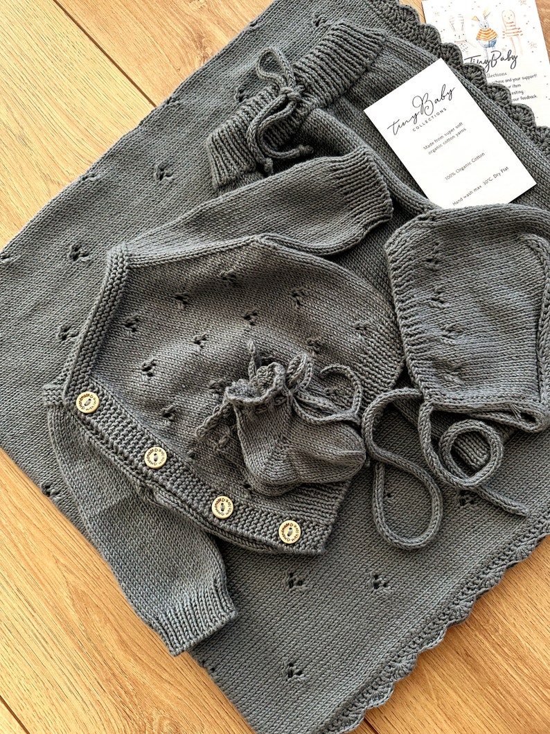K&ouml;nnte beinhalten: Ein graues gestricktes Baby-Outfit-Set mit Cardigan, M&uuml;tze und Booties. Das Set verf&uuml;gt &uuml;ber ein zartes Lochmuster und Holzkn&ouml;pfe. Das Outfit ist aus 100% Bio-Baumwolle gefertigt und nur handwaschbar.