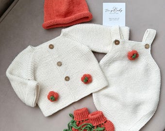 May include: A baby clothing set featuring a knitted orange and green hat, a white cardigan with wooden buttons and flower details, a white romper with flower accents, and orange booties with green vines. The set is made of organic cotton yarn.