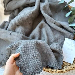 May include: A gray knitted baby blanket with a scalloped edge and small heart-shaped cutouts. The blanket is folded and draped, showing the texture of the knit. The blanket is displayed in a woven basket.