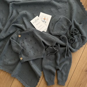 K&ouml;nnte beinhalten: Ein graues Strick-Baby-Outfit-Set bestehend aus M&uuml;tze, Hose und Cardigan. Das Set liegt auf einer grauen Strickdecke mit gewelltem Rand.