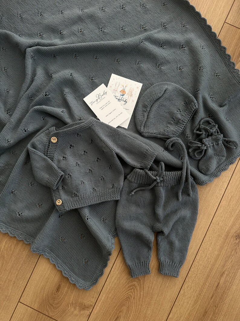 K&ouml;nnte beinhalten: Ein graues Strick-Baby-Outfit-Set bestehend aus M&uuml;tze, Hose und Cardigan. Das Set liegt auf einer grauen Strickdecke mit gewelltem Rand.