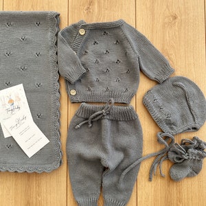 K&ouml;nnte beinhalten: Ein graues Strick-Baby-Outfit-Set bestehend aus M&uuml;tze, Stiefeln, Hose und Cardigan. Das Set ist aus weicher, atmungsaktiver Baumwolle gefertigt und perfekt, um das Baby warm und gem&uuml;tlich zu halten.