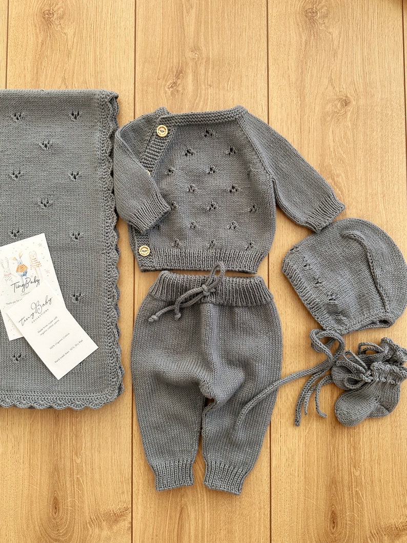 K&ouml;nnte beinhalten: Ein graues Strick-Baby-Outfit-Set bestehend aus M&uuml;tze, Stiefeln, Hose und Cardigan. Das Set ist aus weicher, atmungsaktiver Baumwolle gefertigt und perfekt, um das Baby warm und gem&uuml;tlich zu halten.