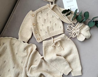 Könnte beinhalten: Ein gestricktes Baby-Outfit-Set in Cremefarbe. Das Set beinhaltet eine Mütze, einen Cardigan, eine Hose und eine Decke. Das Outfit ist mit einem zarten Lochmuster gearbeitet.