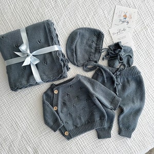 K&ouml;nnte beinhalten: Ein Baby-Outfit-Set in hellblau, bestehend aus einer Strickjacke, einer Hose, einer M&uuml;tze und einer Decke. Das Set ist mit kleinen bl&uuml;tenartigen Mustern verziert und die Strickjacke hat Holzkn&ouml;pfe. Die Decke ist mit einem blauen Band gebunden.
