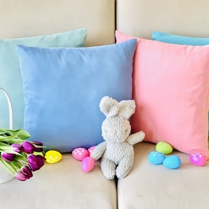 May include: A light blue, a pink, and a light turquoise throw pillow on a beige couch with a gray bunny toy and colorful Easter eggs.