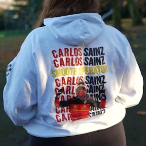May include: White hoodie with a graphic print of a race car driver wearing a red racing suit and helmet. The text "Carlos Sainz" is repeated multiple times on the hoodie.