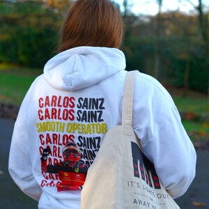 May include: A white hoodie with the text "Carlos Sainz" repeated multiple times and a graphic of a race car driver. The hoodie is being worn by a person carrying a white tote bag with the text "It's lights out and away we go".