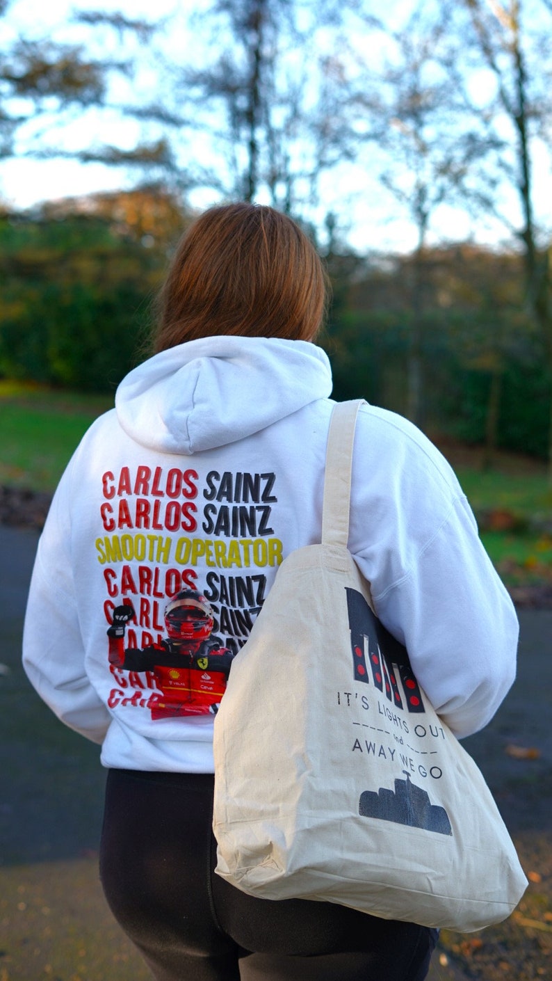 May include: A white hoodie with the text "Carlos Sainz" repeated multiple times and a graphic of a race car driver. The hoodie is being worn by a person carrying a white tote bag with the text "It's lights out and away we go".