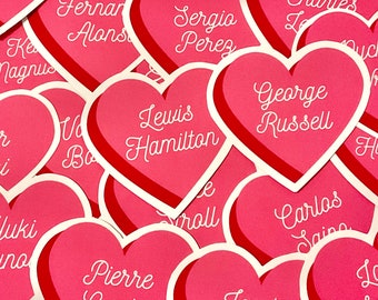 May include: A collection of pink heart-shaped stickers with the names of Formula 1 drivers written in white cursive. The names include Lewis Hamilton, George Russell, Fernando Alonso, Sergio Perez, Carlos Sainz, Yuki Tsunoda, Pierre Gasly, Oscar Piastri, Kevin Magnussen, Nico Hulkenberg, Lance Stroll, and Logan Sargeant.