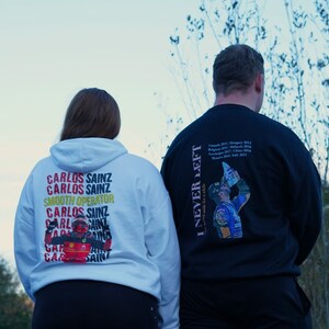 May include: Two people wearing white and black hoodies with a graphic print of a race car driver. The white hoodie has the text "Carlos Sainz" repeated multiple times and the black hoodie has the text "I Never Left" and a list of race wins.
