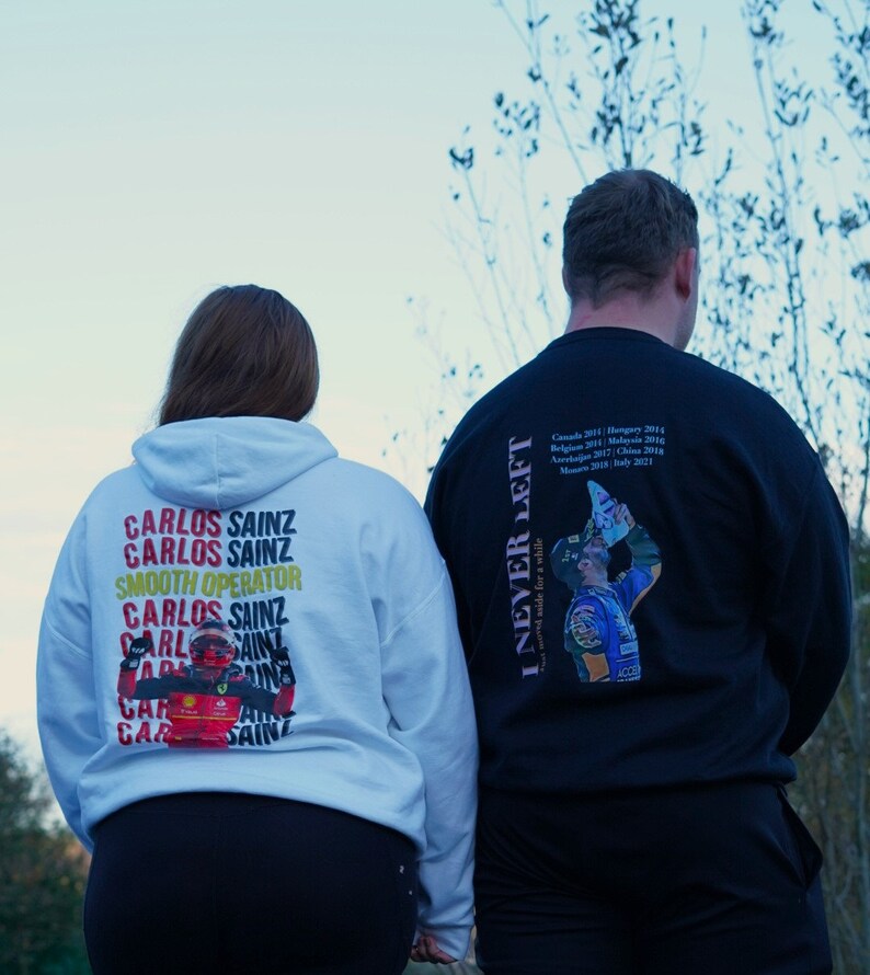 May include: Two people wearing white and black hoodies with a graphic print of a race car driver. The white hoodie has the text "Carlos Sainz" repeated multiple times and the black hoodie has the text "I Never Left" and a list of race wins.