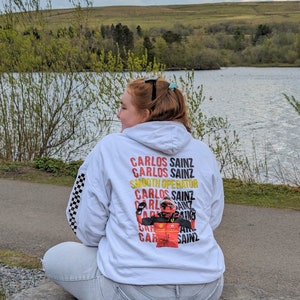 May include: White hoodie with a checkered pattern on the sleeves. The back of the hoodie features a graphic of a race car driver with the text "Carlos Sainz" repeated multiple times.