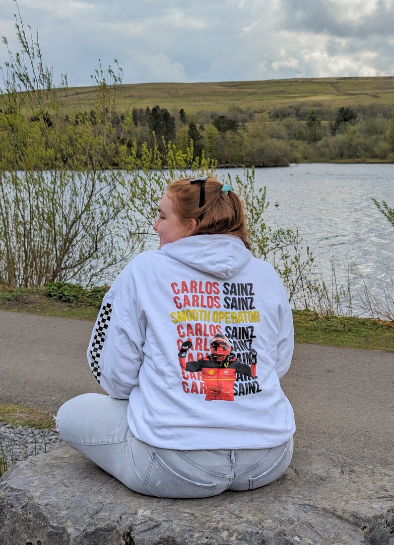 May include: White hoodie with a checkered pattern on the sleeves. The back of the hoodie features a graphic of a race car driver with the text "Carlos Sainz" repeated multiple times.