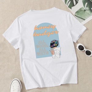 May include: White t-shirt with a blue and white graphic of a person wearing a helmet with the text "honey badger" and "ALL GOOD, ALL WAYS" printed on the back.