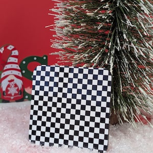 May include: A square coaster featuring a black and white chequered design. The coaster is set in a festive holiday display, with a small Christmas tree and a red backdrop.