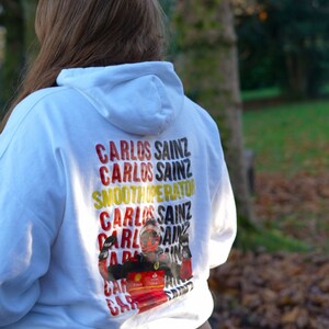 May include: White hoodie with a graphic print of a race car driver with the text "Carlos Sainz" repeated multiple times in yellow, red, and black.