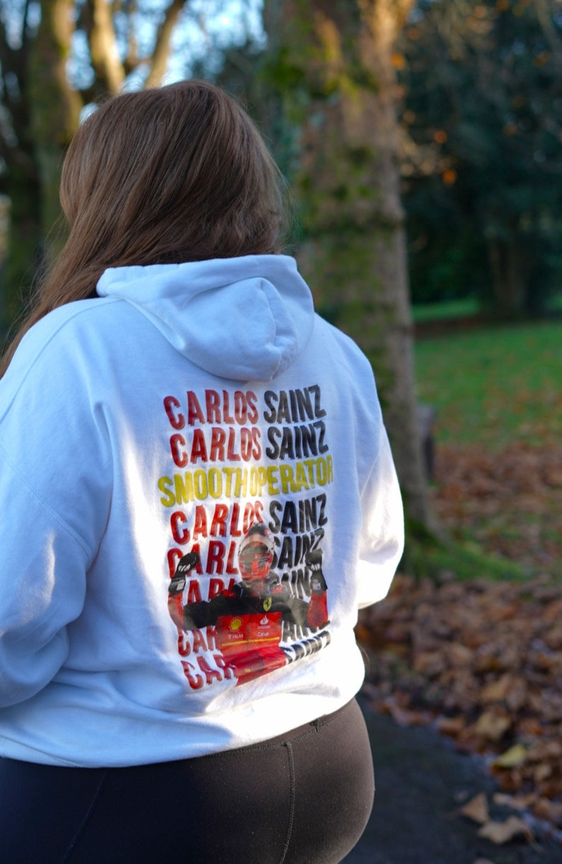May include: White hoodie with a graphic print of a race car driver with the text "Carlos Sainz" repeated multiple times in yellow, red, and black.