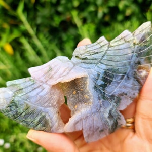 May include: A carved green and purple agate butterfly with a hollow center. The butterfly is carved with intricate details and has a natural crystal formation.