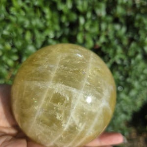 May include: A large, yellow-green crystal sphere with white striations. The sphere is smooth and polished, and it is being held in a hand.