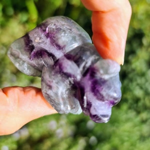 May include: A purple and green fluorite crystal carved into the shape of a bear. The crystal is translucent and has a variety of shades of purple and green.