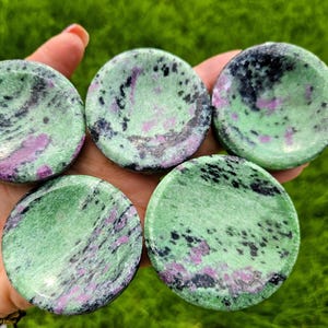 May include: Five round, polished ruby zoisite stones. Each stone has a light green base colour with black and pink speckles. The stones are held in a hand against a blurred green background.