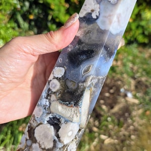 May include: A rectangular crystal obelisk with a polished surface. The stone features a mix of colours, including white, grey, blue, and brown, with a marbled pattern. The obelisk is held in a hand against a blurred green background.