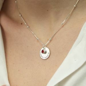 Puede incluir: Un collar de plata con un colgante redondo que dice "79.074°W 43.6532°N" y tiene una pequeña piedra preciosa roja. El collar tiene una cadena de bola de caja.