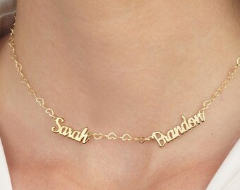 May include: A gold-colored necklace with the names "Sarah" and "Brandon" in cursive script. Small heart-shaped charms connect the names and form the necklace chain. The necklace is worn against a white shirt.