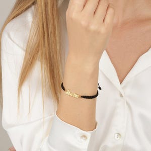 May include: A black braided bracelet with a gold-colored nameplate that reads "Justin" in a stylized font. The bracelet is worn on a wrist, and the background is a white shirt.