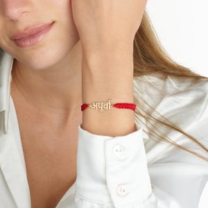 Puede incluir: Una pulsera roja tejida con un dije dorado que deletrea una palabra en una escritura. La pulsera se usa en una muñeca y el fondo es una camisa blanca.