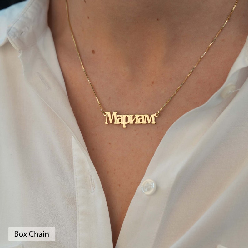 May include: A gold chain necklace with a nameplate pendant that reads "Mariam" in a bold, blocky font. The necklace is on a model wearing a white shirt.