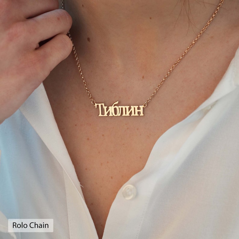May include: A gold necklace with a nameplate pendant that reads "Тиблин". The necklace is on a rolo chain.