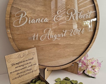 May include: A wooden guest book box with a clear glass lid and a wooden base. The lid has the text "Bianca & Robert 11 August 2024" written on it. The box is filled with small green wooden hearts.